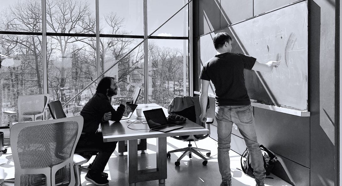 A man sits at a desk in profile in a sunny room with large windows. Another man makes calculations on a chalkboard.